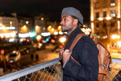 Young man in a wool cap and backpack stands on a bridge at night, gazing at the illuminated paris