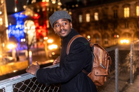 Young black man wearing a coat and a beret, carrying a backpack, standing on a bridge overlooking