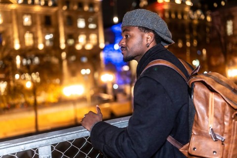 Young black man wearing a cap and coat with a brown backpack, standing on a bridge, thoughtfully