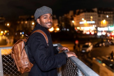 Young black man smiling while looking at the viewer, standing on a bridge overlooking the