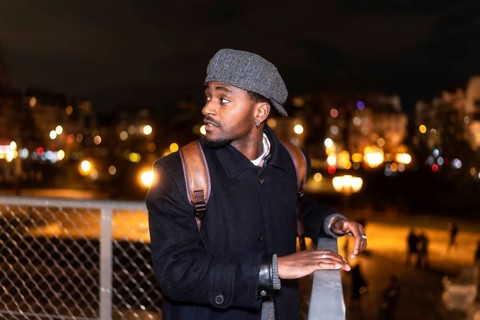 Young man in a flat cap and backpack gazes across a paris bridge at blurred city lights and bokeh