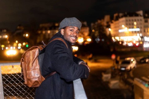 Cheerful male tourist wearing a classic cap and brown backpack, confidently posing on a bridge rail