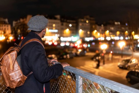 Young black man wearing a cap and backpack stands on a bridge, overlooking a busy parisian street
