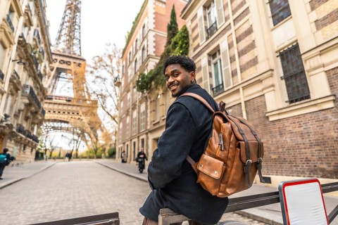 Happy black man tourist with a backpack sitting on a railing on a charming parisian street, smiling