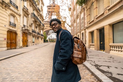 Young african man with glasses and backpack smiles confidently while walking a cobblestone paris