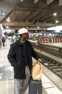 Young man with a baseball cap and glasses standing on a train station platform, holding a shopping