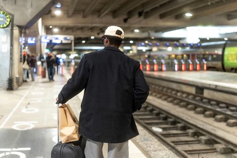 Man standing on a train station platform, seen from behind, pulling a suitcase and carrying a paper