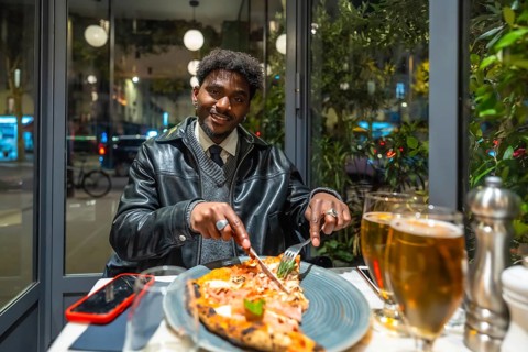 Young man smiling and cutting a delicious pizza slice with a knife and fork, enjoying a relaxing