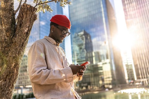 Young black man wearing a red beanie and sunglasses checking his mobile phone outdoors, standing by
