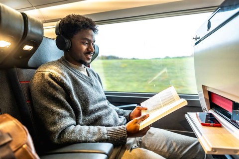 Young man wearing headphones and reading a book while traveling comfortably by high speed train,