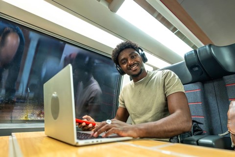 Young black man with headphones smiling while working on a laptop and using a smartphone during a