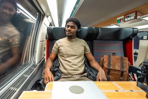 Young black man in headphones and smart casual clothes working on a laptop and gazing out the train