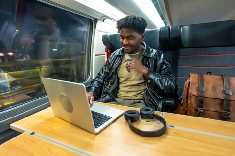 Young man using a laptop and headphones, smiling while traveling by train, reflecting modern
