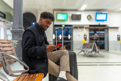 Young black man sitting comfortably on a wooden bench at a train station, using his red smartphone