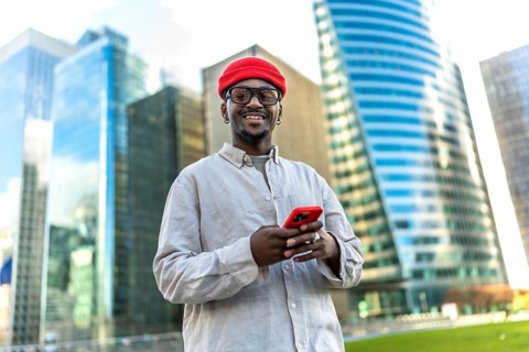 Young black man smiling, looking at the camera while using a smartphone in a modern urban district