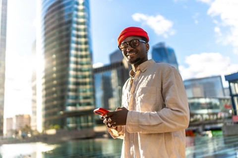 Young man smiling, holding and looking at a mobile phone, standing in a vibrant urban environment