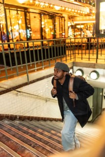 Young black man in beret and coat climbs subway stairs at night, backpack on shoulder, illuminated