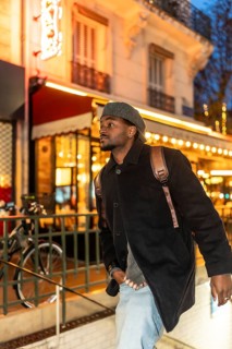 Young black man wearing a beret, coat, and backpack exploring the brightly lit streets of paris,
