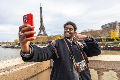 Smiling young man capturing a travel moment with his smartphone, standing on a bridge overlooking