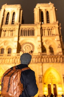 Tourist with backpack and hat seen from behind, gazing at notre dames illuminated gothic facade at