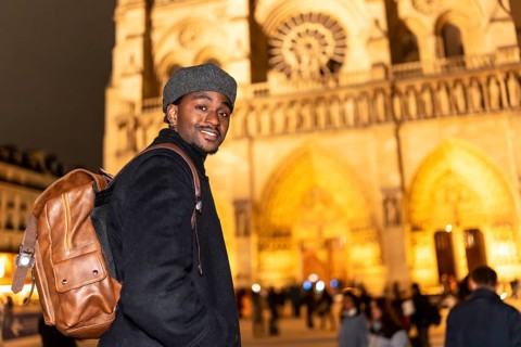 Young black man with a backpack smiling, visiting notre dame cathedral in paris france at night,