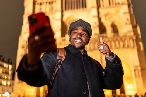 Young man smiling and giving a thumbs up gesture, taking a selfie with his smartphone in front of