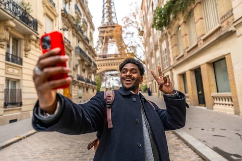 Young man visiting paris, smiling and making a peace sign while taking a selfie with a smartphone,