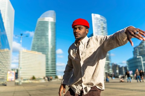 Black man wearing a red beanie and casual clothing moving rhythmically on a sunny day in an urban