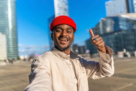 Young black man with a red beanie taking a close up selfie in an urban setting, smiling and giving