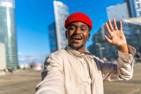 Young black man taking a selfie, smiling broadly and waving at the camera, showing a friendly