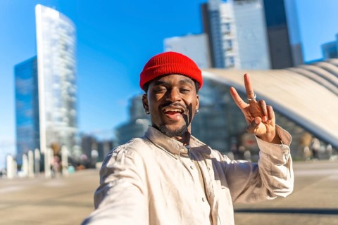 Young black man smiling and making a peace sign gesture while taking a selfie outdoors in a modern
