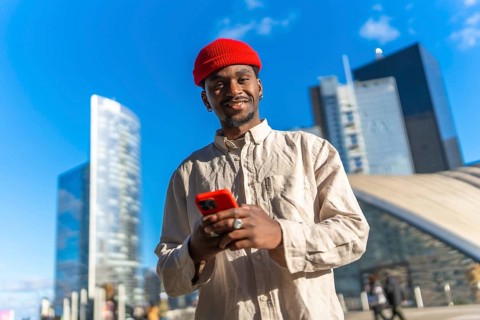 Young black man smiling at camera while using a red smartphone outdoors, standing in a contemporary