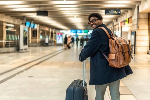 Smiling african man carrying backpack and pulling luggage, looking back while walking through a