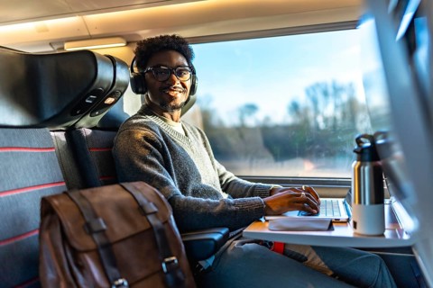 Young black man smiling while traveling on a train, wearing headphones and typing on his laptop,