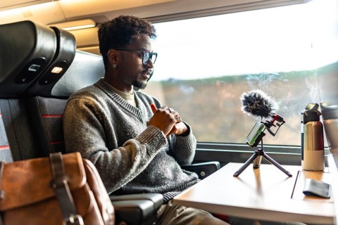 Young black man vlogging on a train, using smartphone and microphone on a tripod during a business