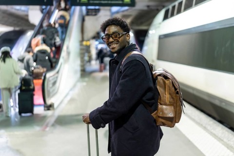 Smiling black man stands on a modern train platform with suitcase and backpack, ready for a