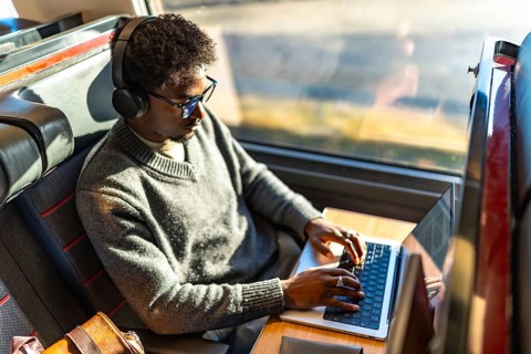 Young black man focused on laptop on a train, wearing headphones and glasses while working remotely