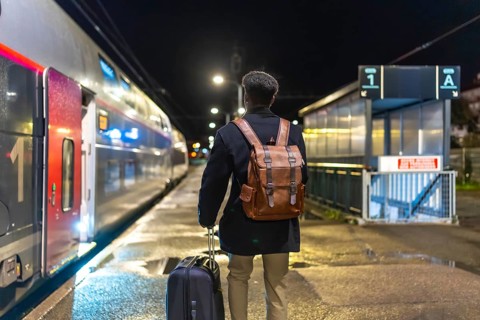 Man in coat and backpack pulls rolling luggage on a dim train platform at night, boarding for a