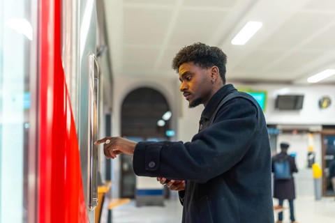 Young black man standing at a ticket machine in a modern train station, purchasing a ticket for his