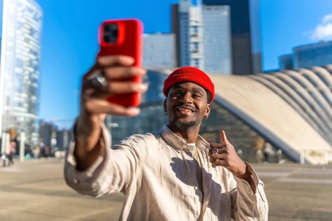 Young black man smiling while taking a selfie with a red smartphone and giving a thumbs up gesture,
