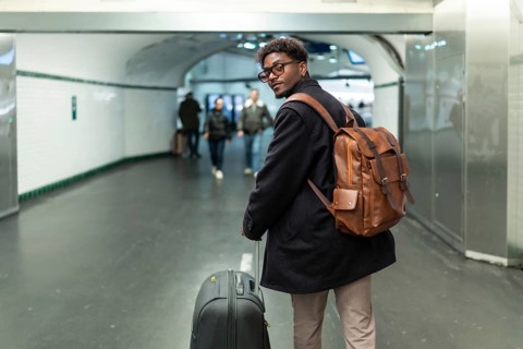 Young man with a stylish leather backpack and rolling luggage is pausing to look back in a modern