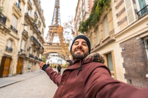 Happy bearded man in winter clothes takes a smiling selfie with the eiffel tower behind him,