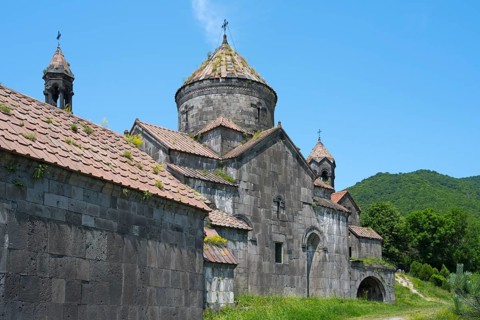 Old monastery building with stone structures and dome in front of a green mountain landscape,