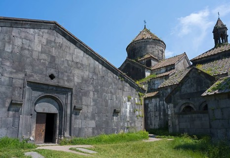 Massive stone church with large windows and an open wooden gate under a blue sky, Haghpat
