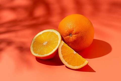 On the coral surface, next to two neatly sliced slices, there is a whole orange. Sunlight casts