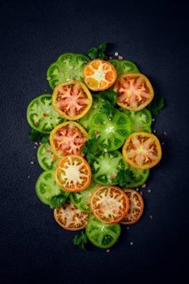 Sliced green tomatoes, on a dark surface, garnished with herbs and salt, top view