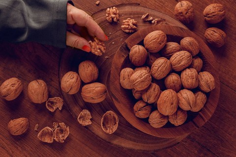 A woman's hand taking a walnut from a wooden bowl filled with walnuts. There are shells and walnuts