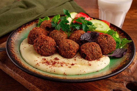 A plate holds falafel balls, tahini, salad with tomatoes and cucumbers, and a drink. The setting is