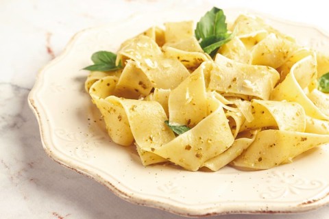 Fresh papardelle pasta, with basil and pesto sauce, is served, on a white plate, with a fork held