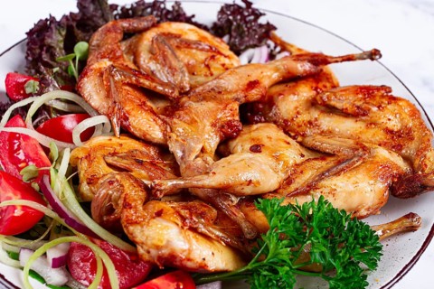Baked quail, served on a plate with crispy lettuce leaves, fresh tomatoes and herbs, on a white
